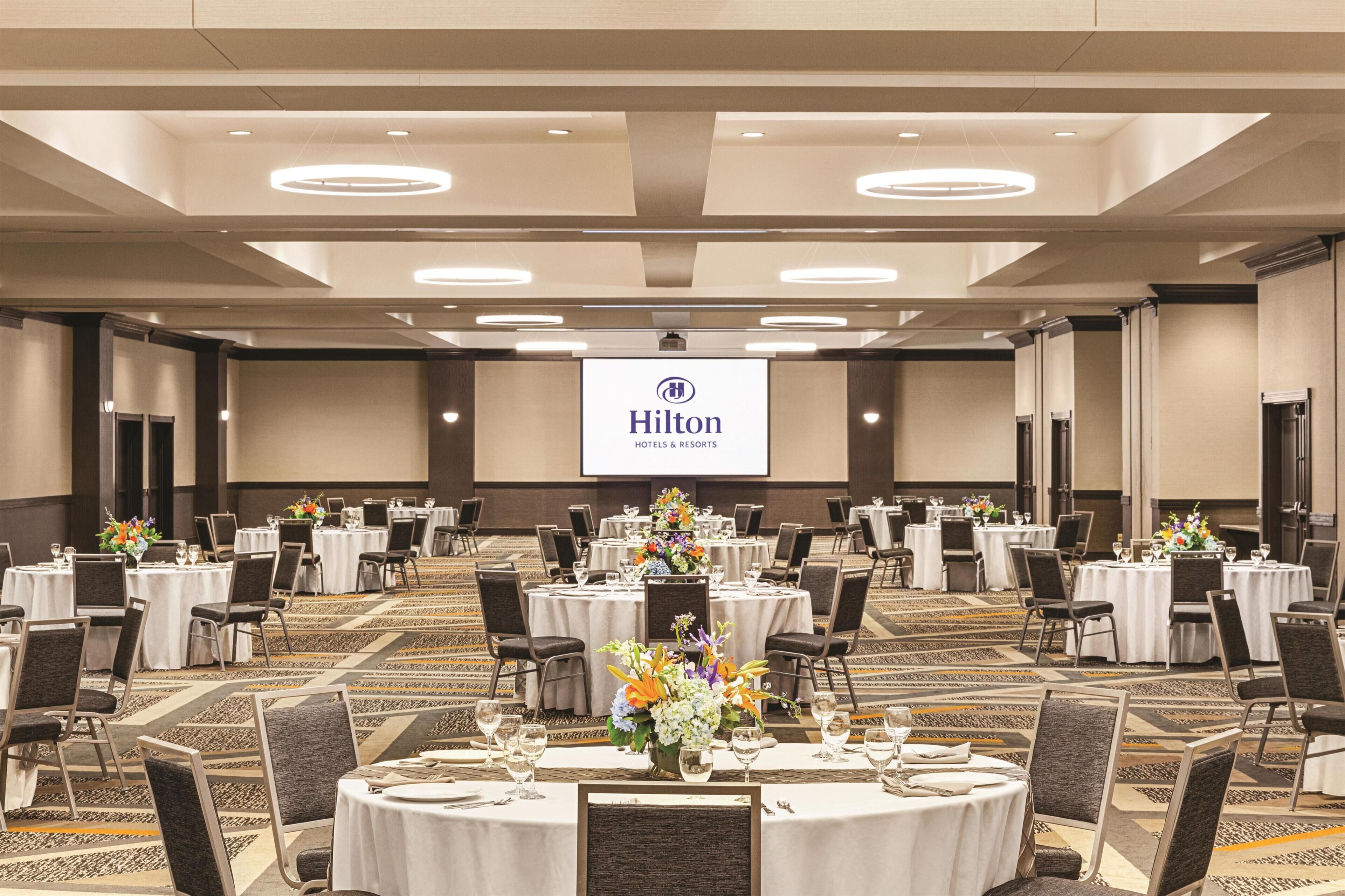 Un amplio salón de conferencias con varias mesas, sillas y una pantalla para presentaciones que muestra el logotipo de Hilton.
