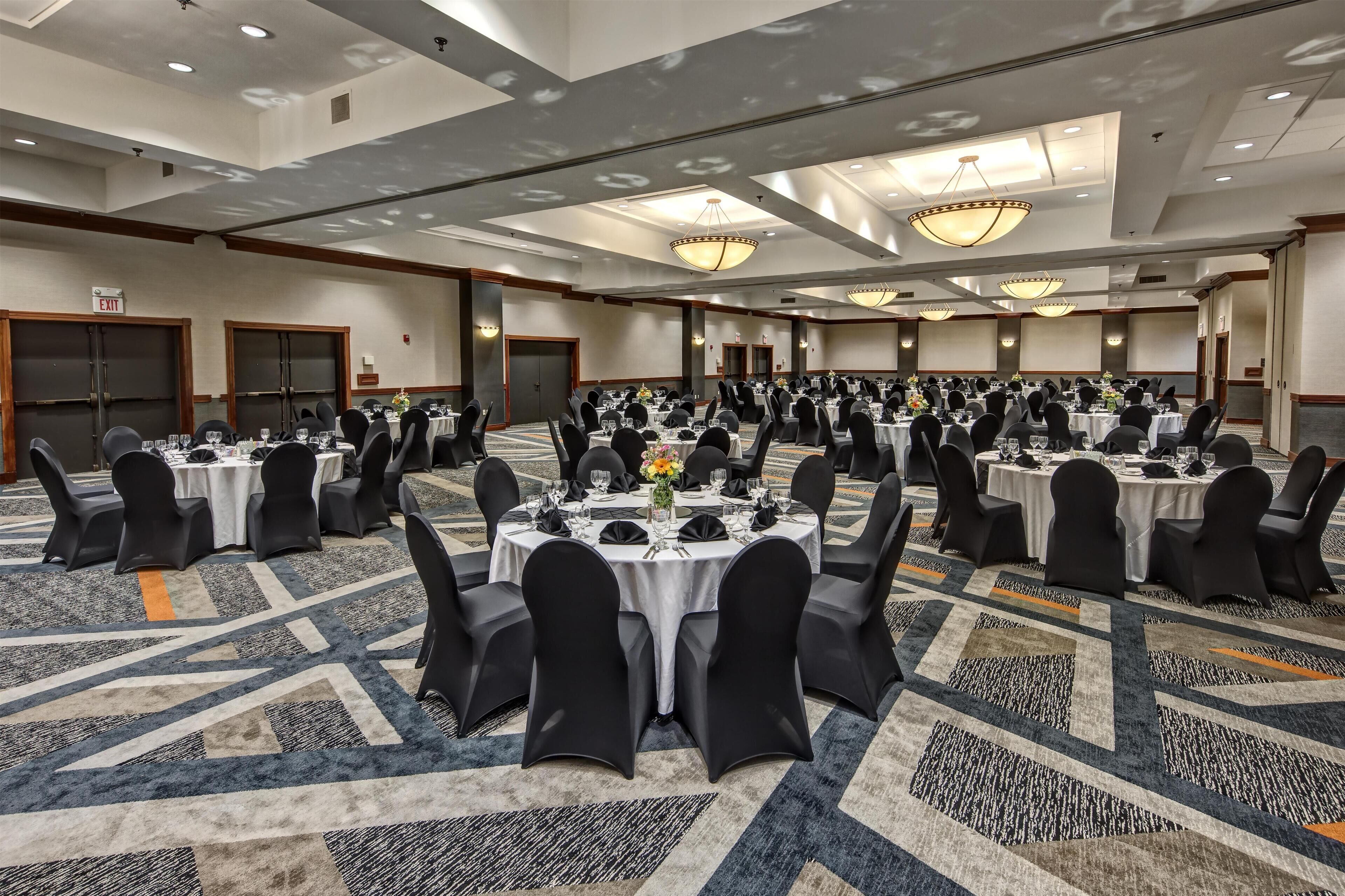 A large banquet hall with multiple round tables, black chairs, and chandeliers.