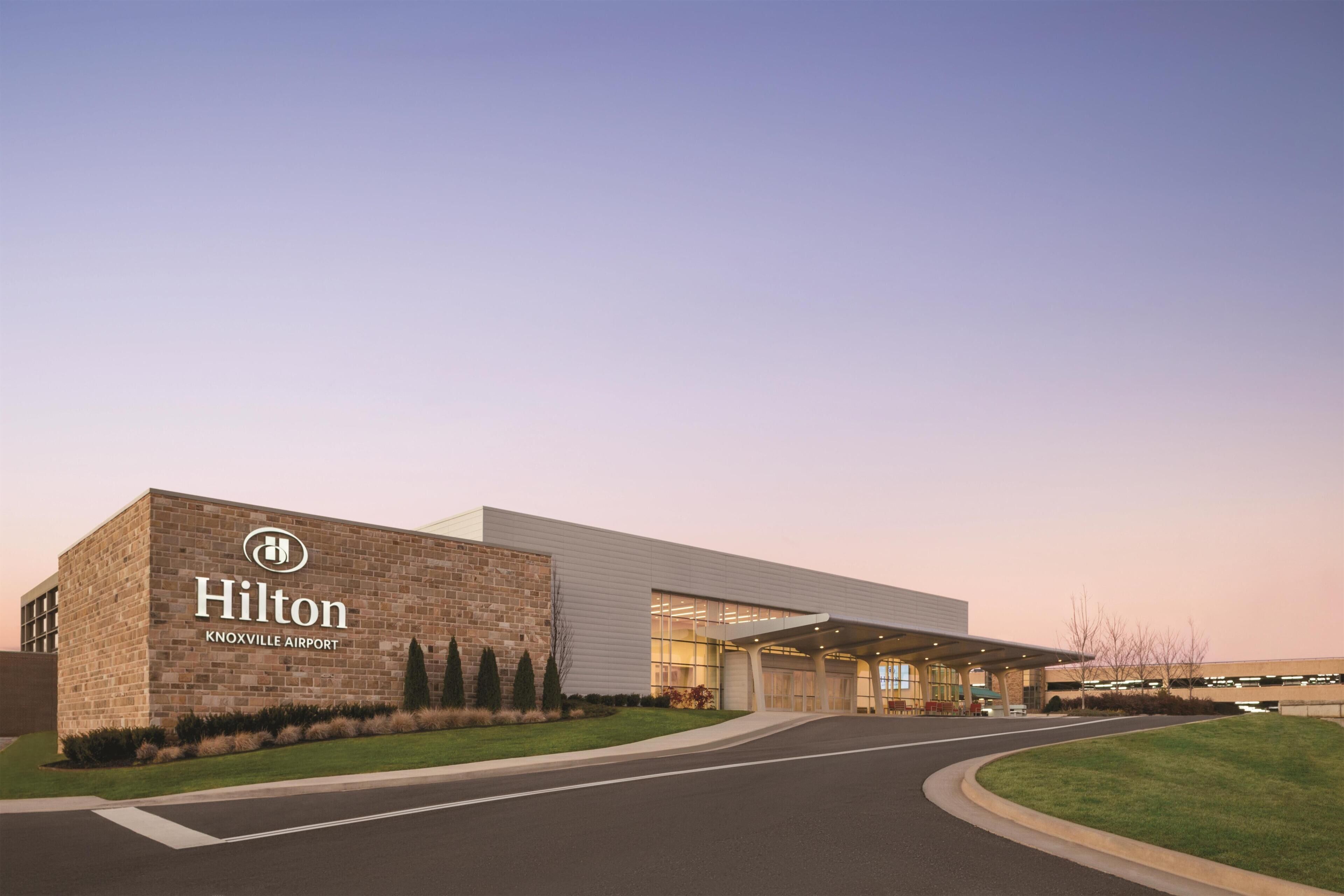 A modern building with a 'Hilton Knoxville Airport' sign, a covered entrance, and a landscaped area with trees.