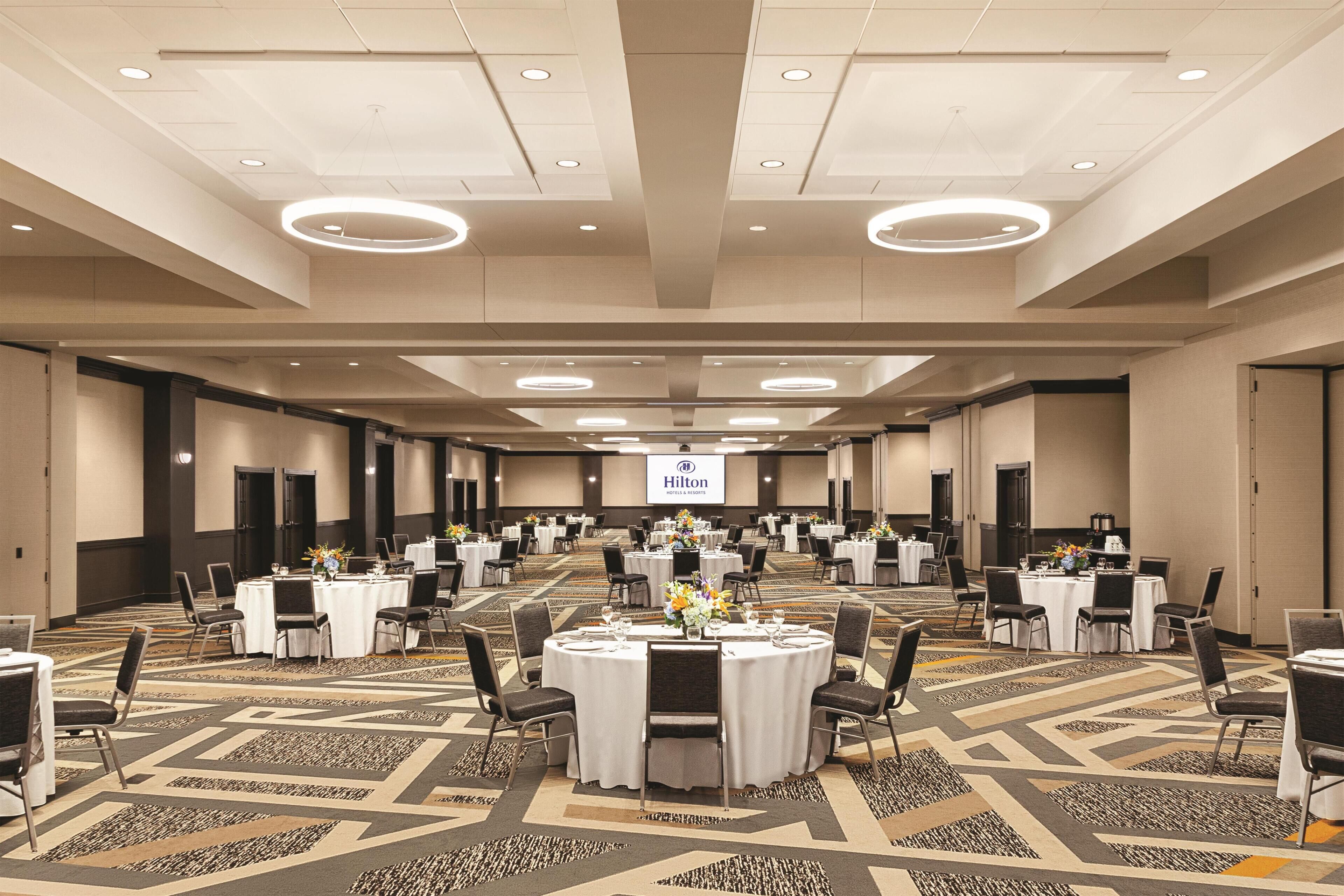Un salón de conferencias amplio con mesas redondas, sillas y una pantalla proyectora mostrando 'Hilton'.