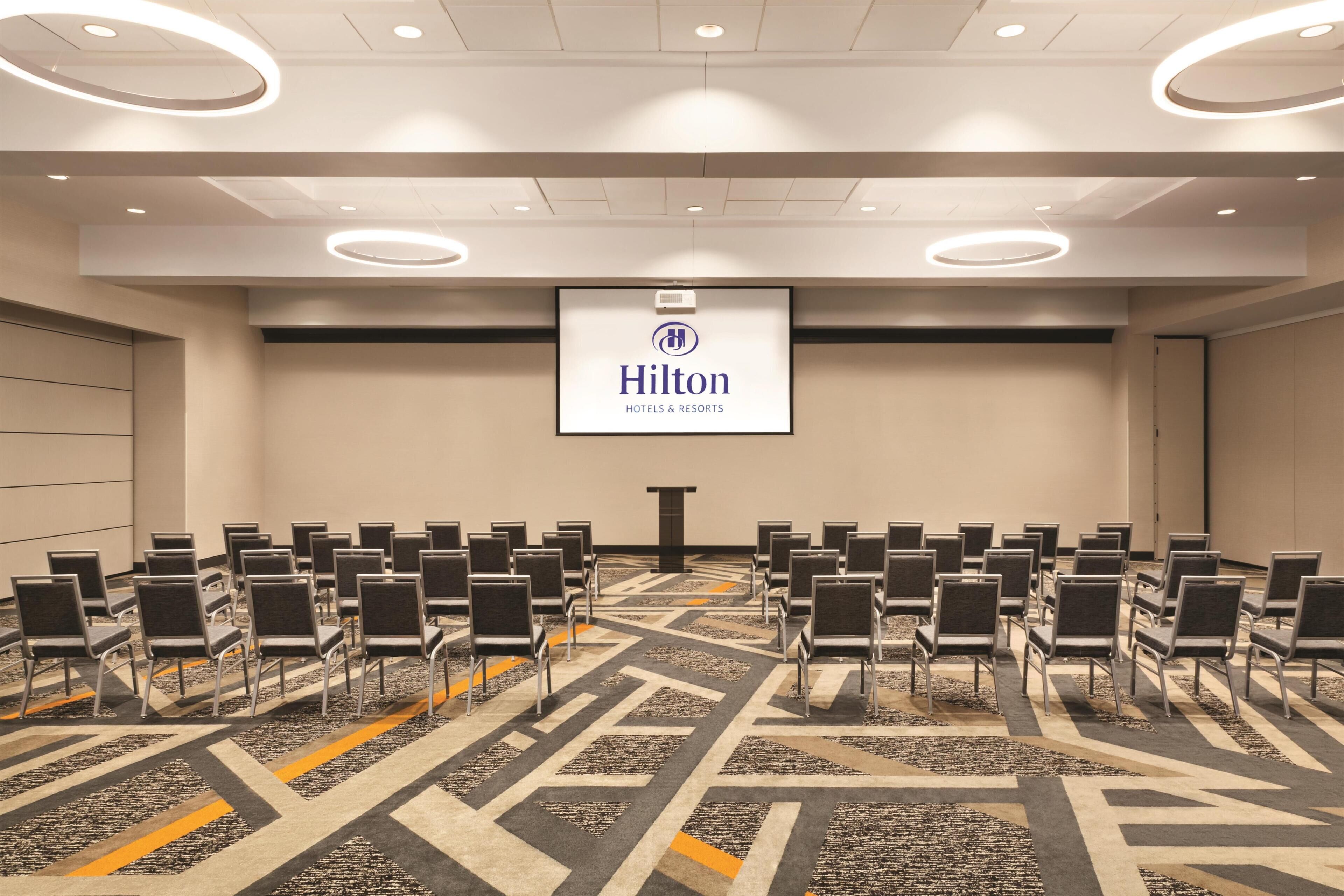 A conference room with rows of chairs, a podium, and a Hilton Hotels & Resorts logo on the wall.