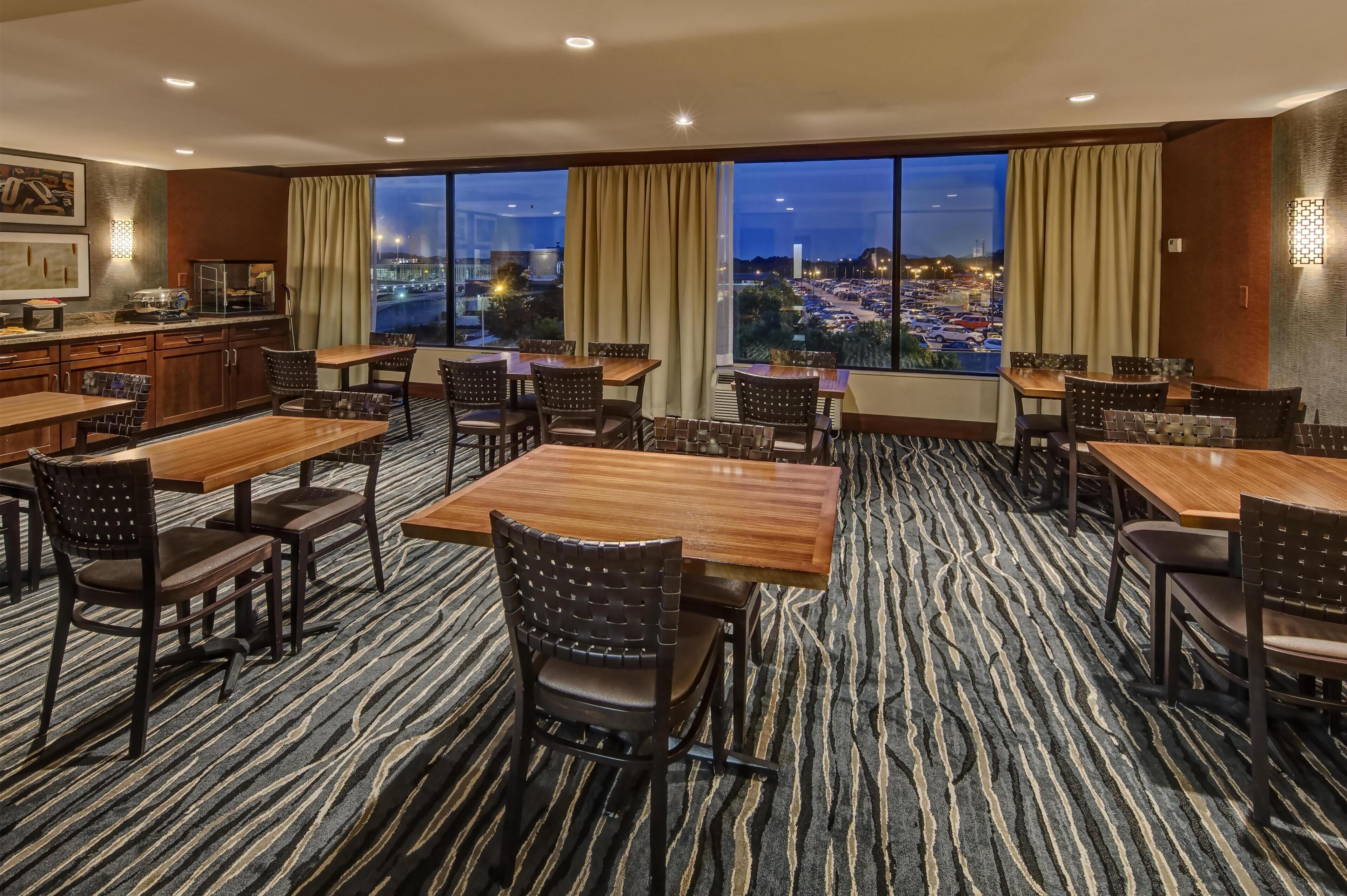 A hotel dining area with multiple tables and chairs, large windows with curtains, and a view of a cityscape at dusk.