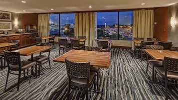 A hotel dining area with multiple tables and chairs, large windows with curtains, and a view of a cityscape at dusk.