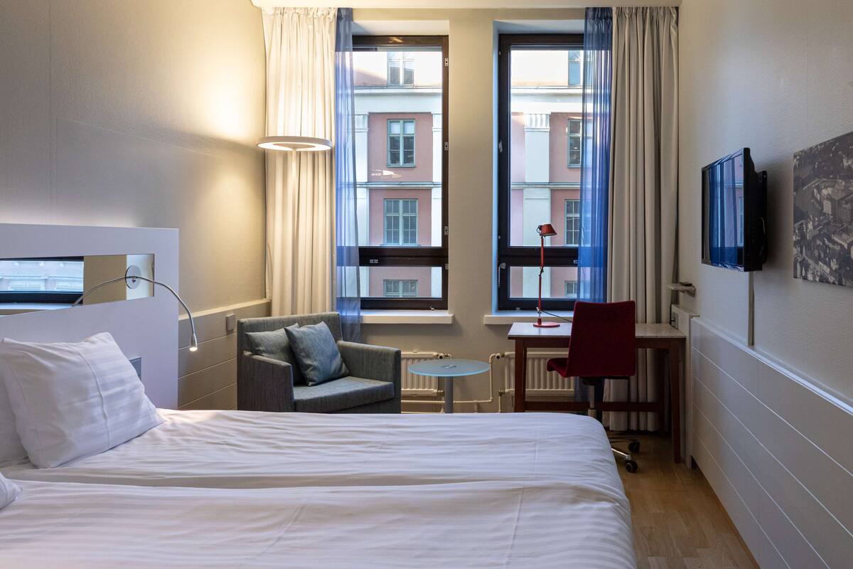 Quarto de hotel com cama, escrivaninha, cadeira e televisão.