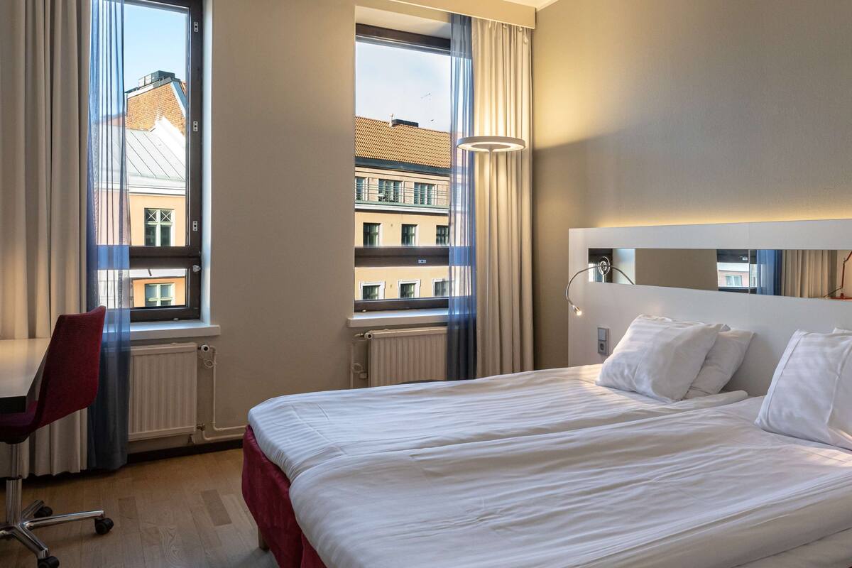 Quarto de hotel com uma cama, uma mesa e uma cadeira. Há janelas com cortinas, um radiador e um ponto de luz fixado na parede.