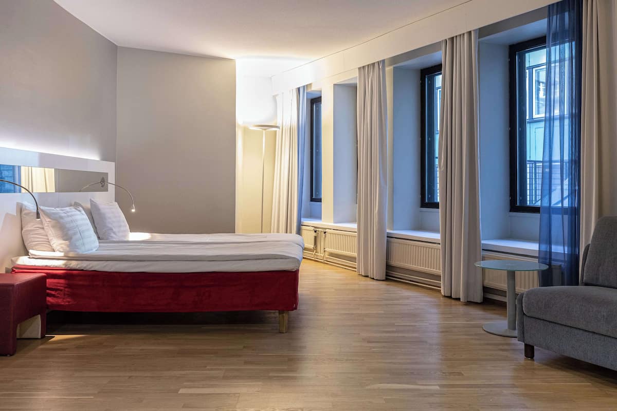 Quarto de hotel moderno com cama, um banco estofado vermelho, sofá cinzento e janelas grandes com cortinas.
