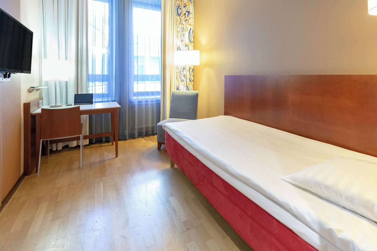 Quarto de hotel com cama, escrivaninha, cadeira, televisão e janela com cortinas.