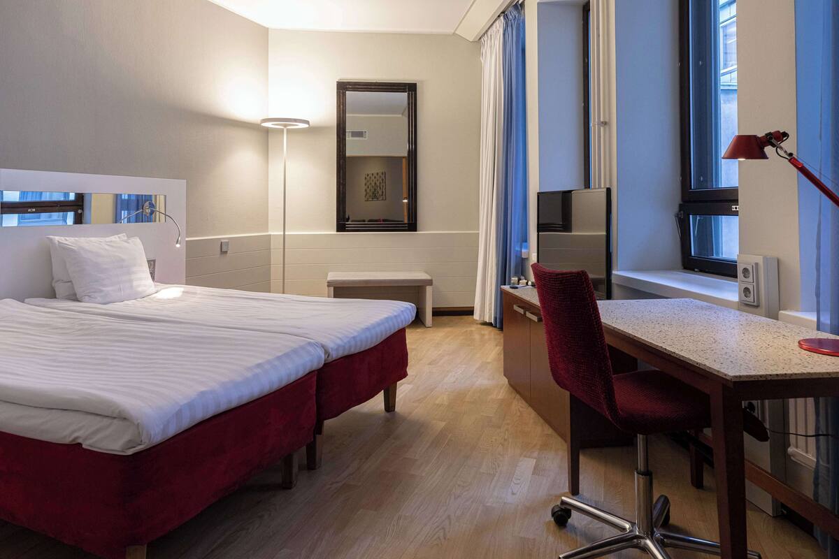 Quarto de hotel com duas camas, uma escrivaninha, uma cadeira, televisão e uma janela grande com cortinas.