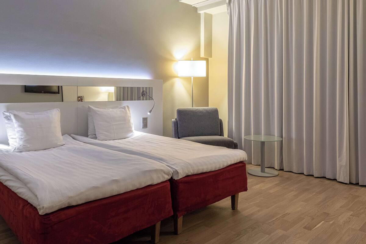 Quarto de hotel com duas camas, uma cadeira, uma mesinha e um abajur.