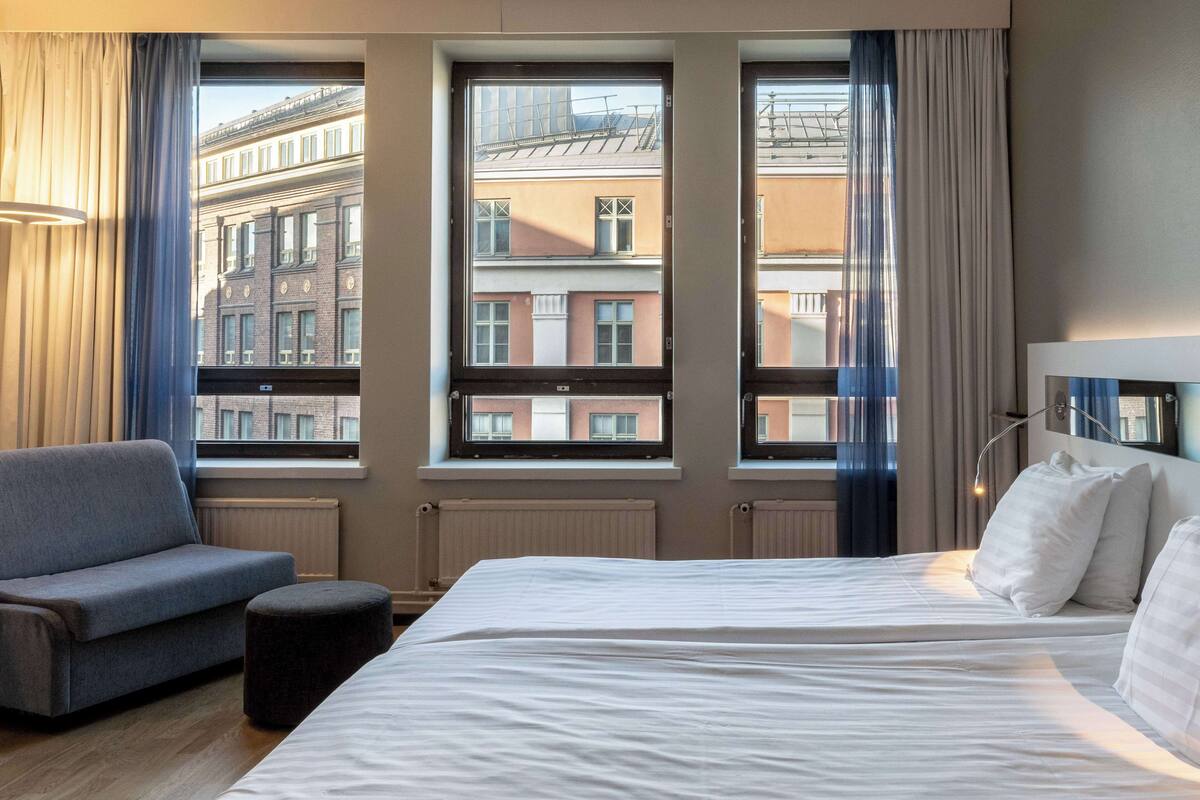 Quarto de hotel com uma cama grande, um sofá e três janelas com vista para edifícios.