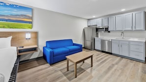 Une chambre d’hôtel moderne avec un lit, un canapé bleu, une table en bois, un coin cuisine avec des placards et des appareils électroménagers, et un tableau représentant un paysage accroché au mur.