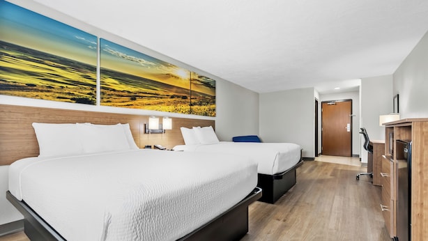 Une chambre d’hôtel avec deux lits, un bureau, une chaise, une armoire et un grand tableau représentant un paysage accroché au mur.