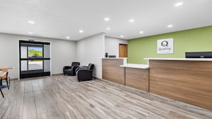 Un espace d’accueil d’hôtel avec un comptoir de réception en bois, un mur végétal orné d’un panneau indiquant « Qualité », un coin salon avec des chaises, et une vue à travers une porte vitrée.