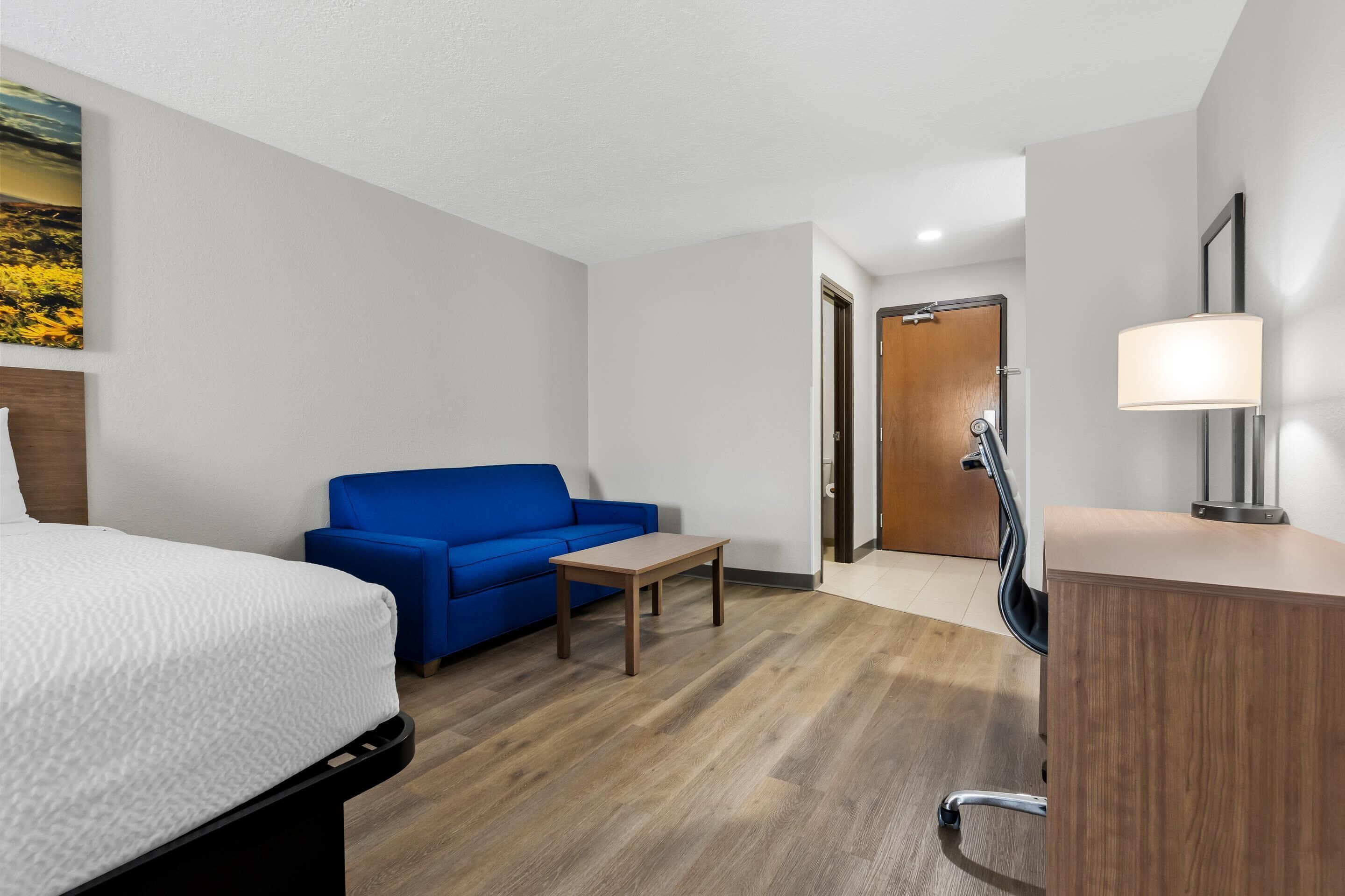 A hotel room with a bed, a blue sofa, a small table, a desk with a lamp, and a wooden door.