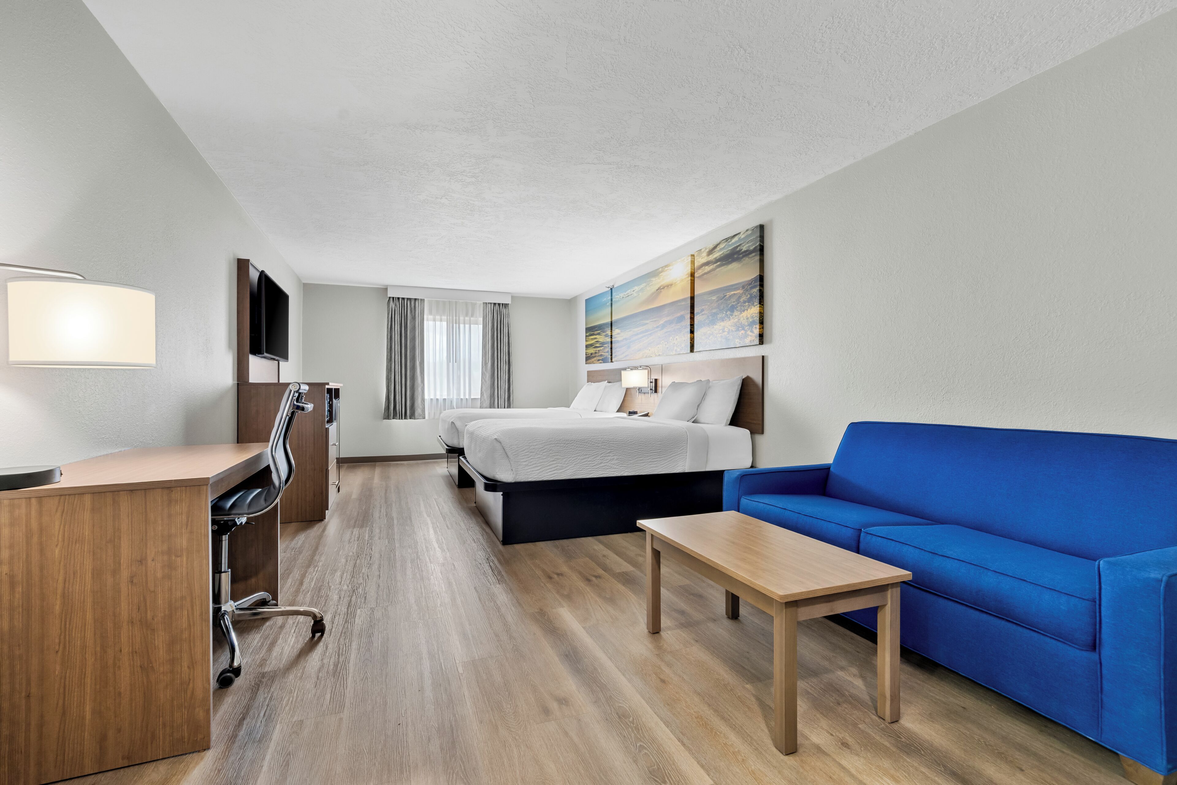 Une chambre d’hôtel avec un bureau, deux lits, un canapé bleu et un tableau au mur.