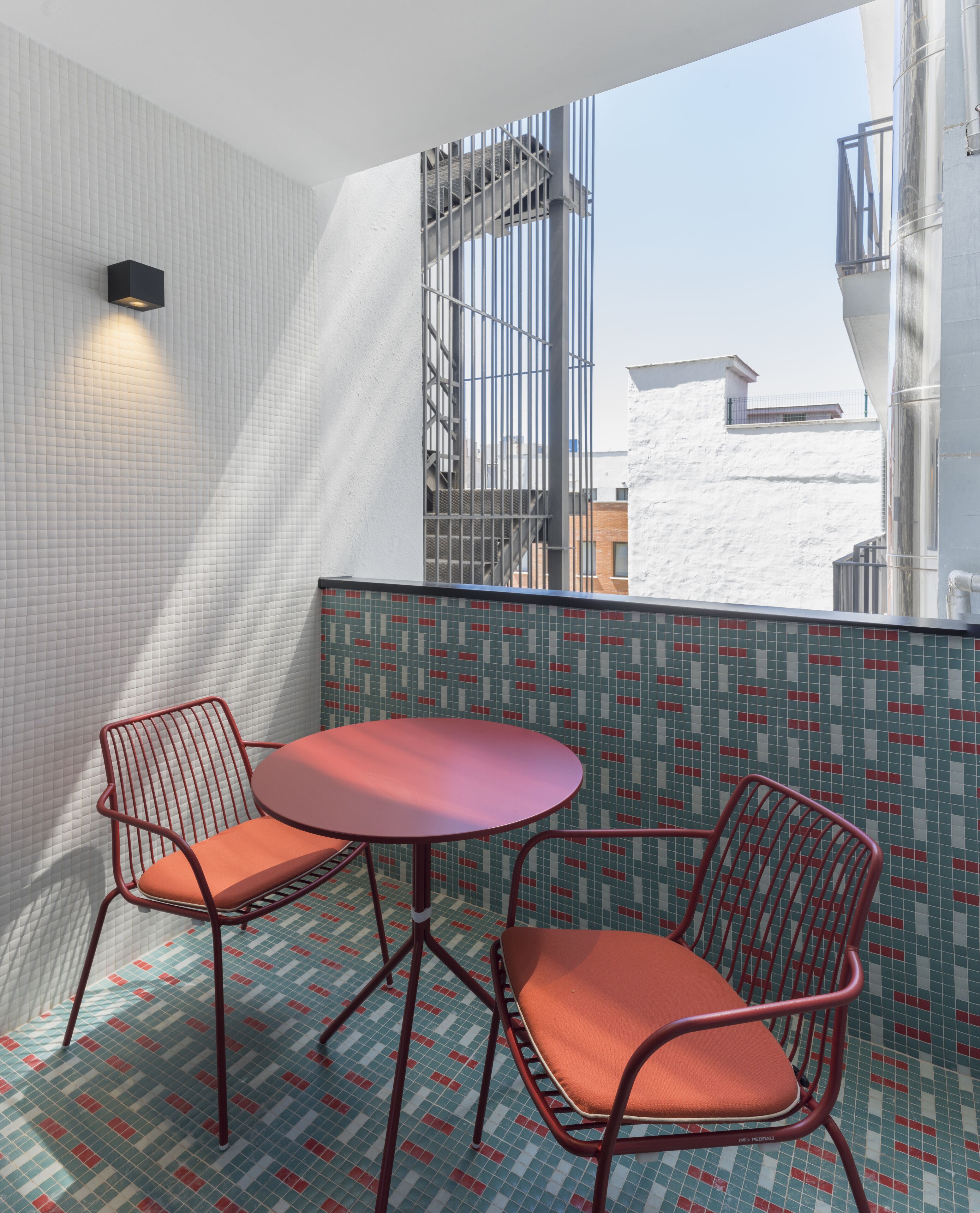 Een klein balkon met twee metalen stoelen en een ronde tafel, een betegelde vloer en uitzicht op gebouwen door een metalen hekwerk.