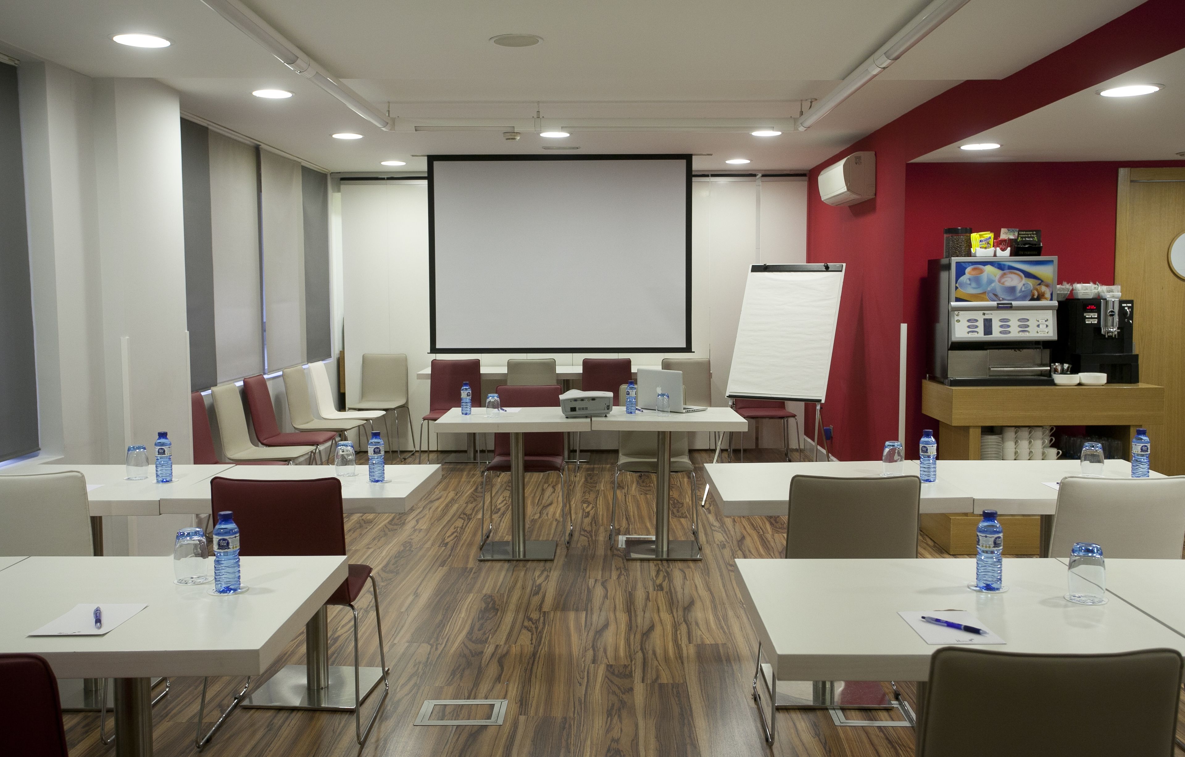 Un salón de conferencias con una pantalla grande, una pizarra y una máquina de café. Las mesas están equipadas con botellas de agua y bolígrafos.