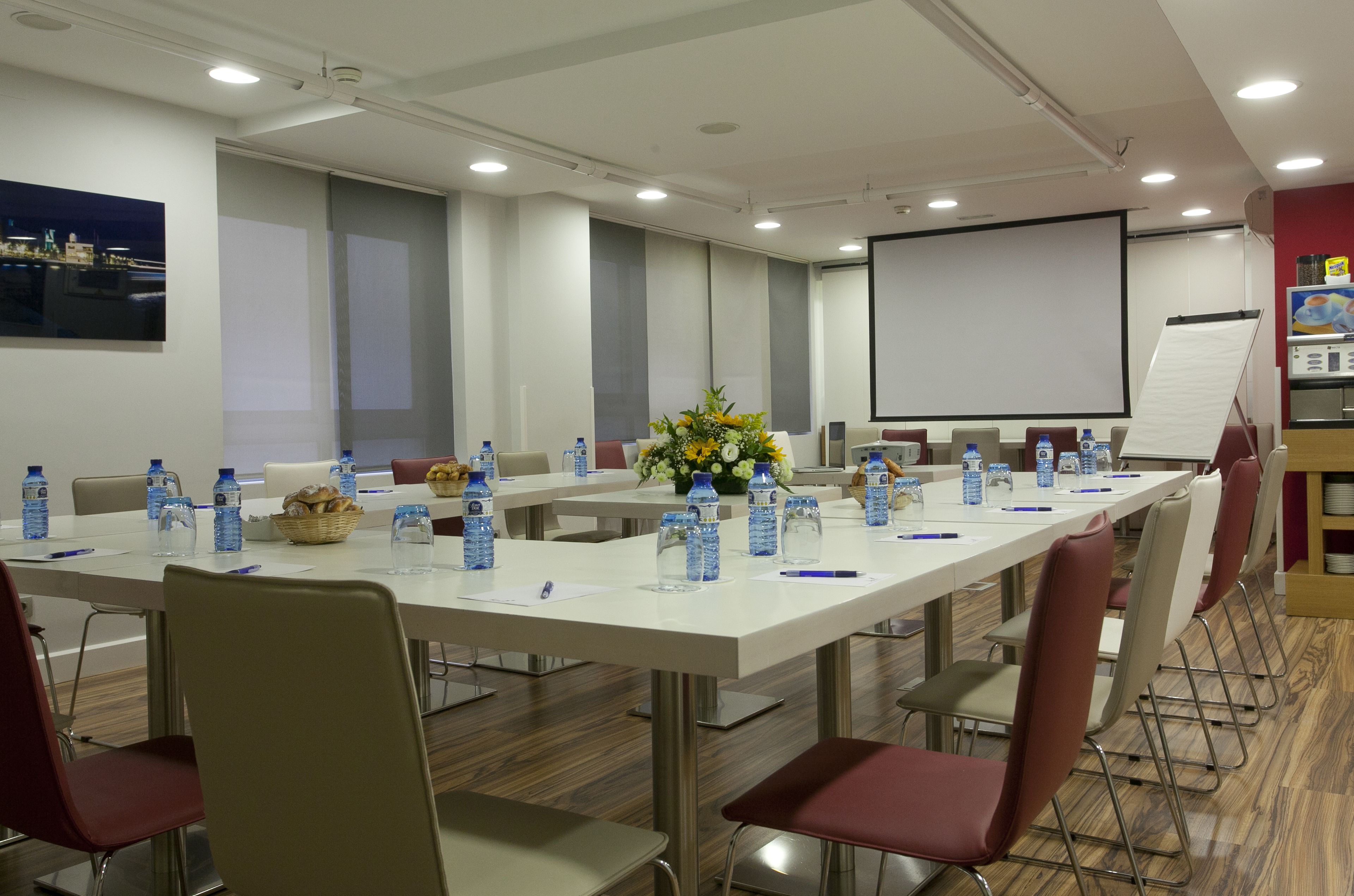 Un salón de conferencias con largas mesas, sillas, botellas de agua, una pantalla de proyector y una pizarra.