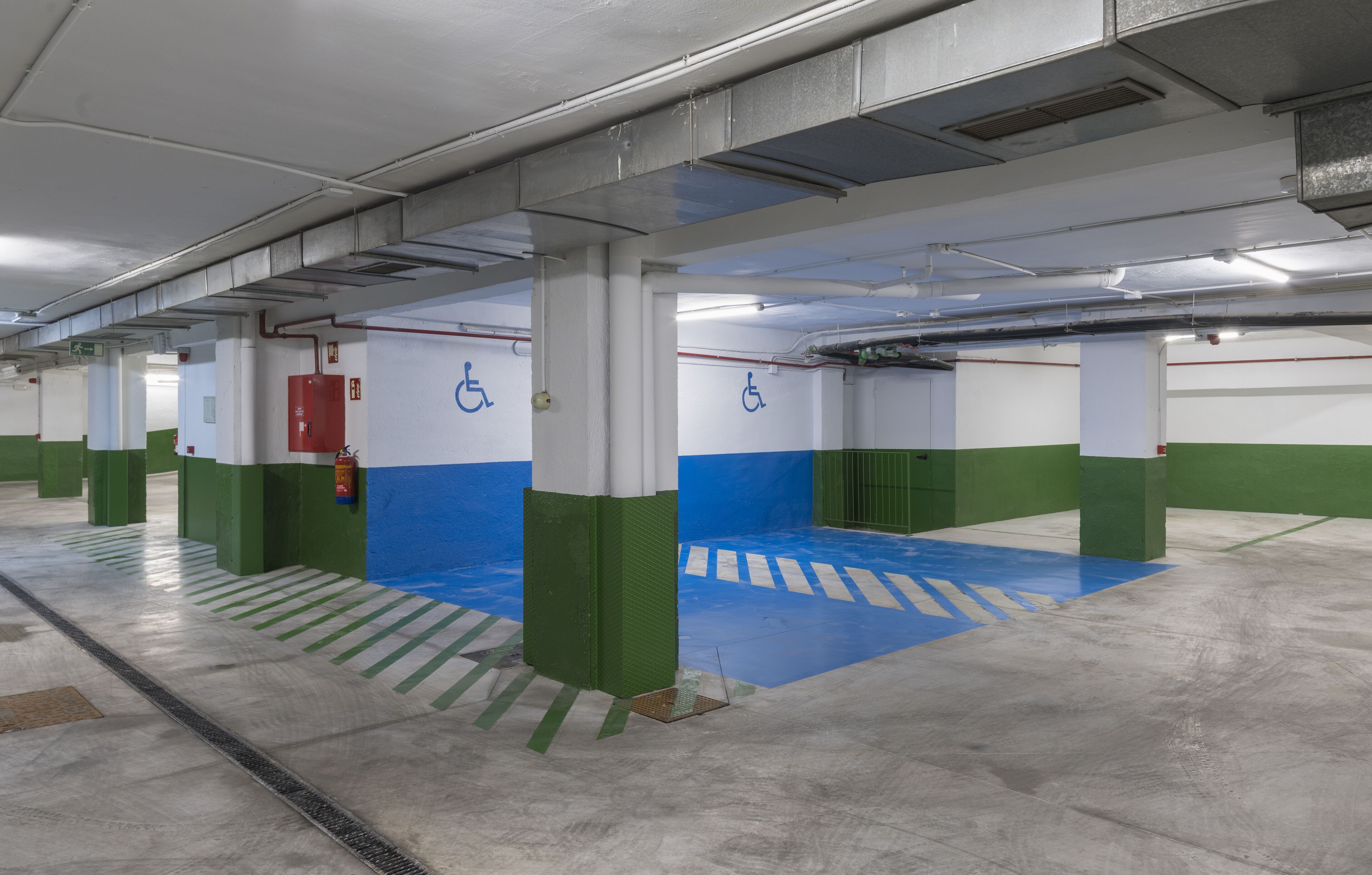 Un aparcamiento de varios niveles con pilares verde y blanco, marcas en el suelo azules y señales de acceso para sillas de ruedas.