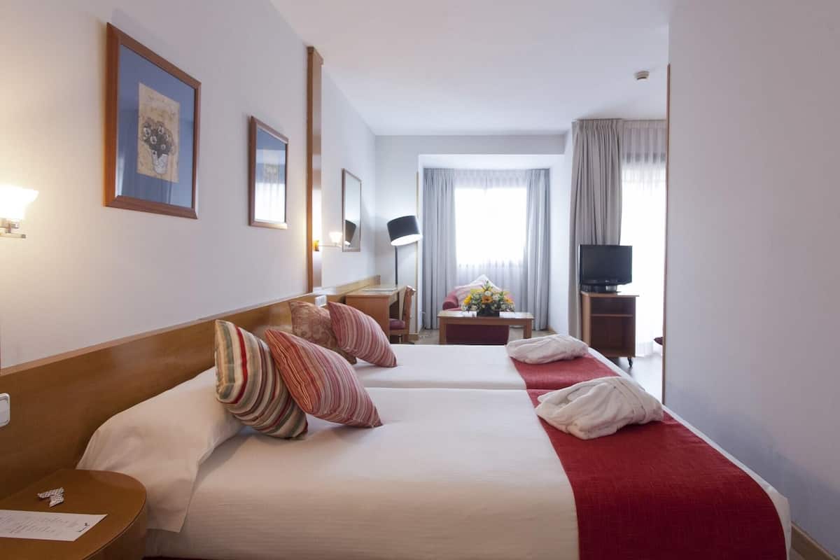 A hotel room with two beds, a desk, a chair, a TV, and a window with curtains.