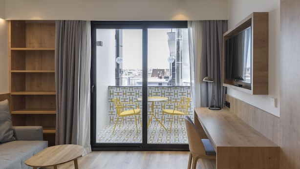 Una habitación de hotel moderna con un sofá, una mesa de madera, un televisor montado en la pared y un balcón con sillas amarillas.