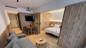 Una habitación de hotel moderna con suelo de madera, un sofá, una mesita redonda, una cama y un televisor fijado a la pared.