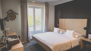 Habitación de hotel con una cama grande, un escritorio con lámpara, una silla y vista a áreas verdes por la ventana.