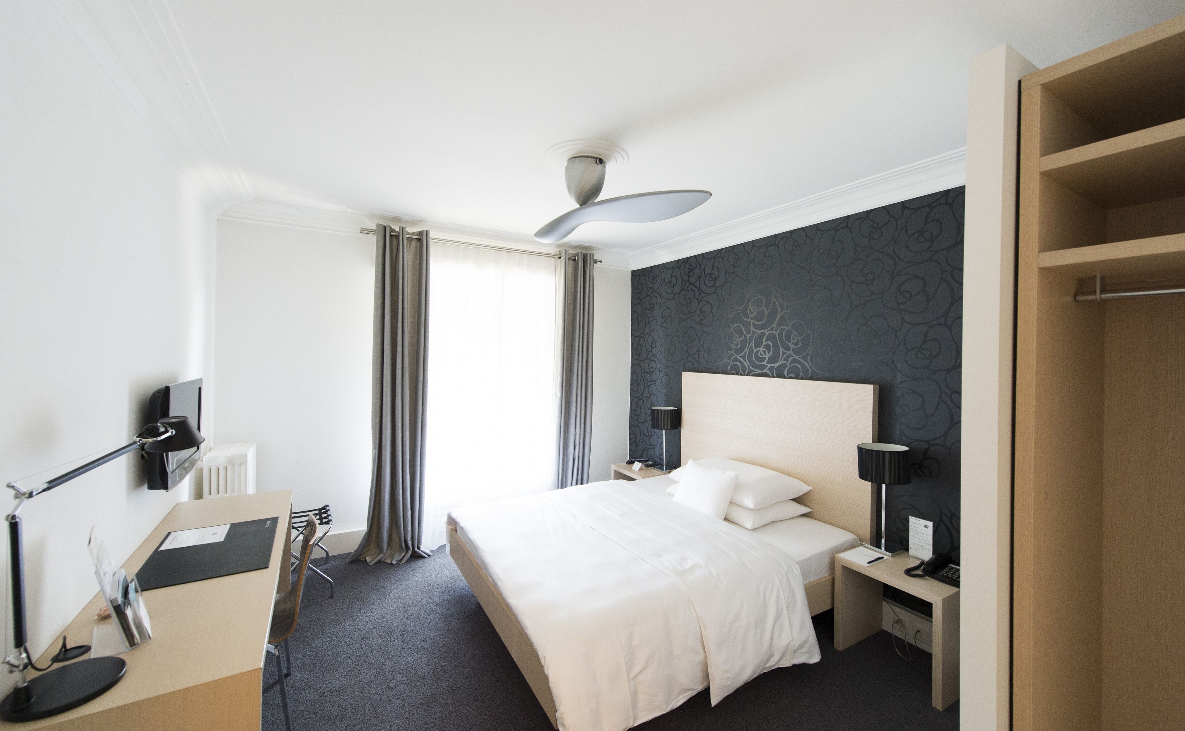 Hotellihuone, jossa on sänky, yöpöydät, työpöytä, tuoli, pöytälamppu, kattotuuletin ja ikkuna, jossa on verhot.