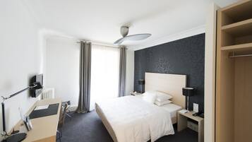 Hotellihuone, jossa on sänky, yöpöydät, työpöytä, tuoli, pöytälamppu, kattotuuletin ja ikkuna, jossa on verhot.