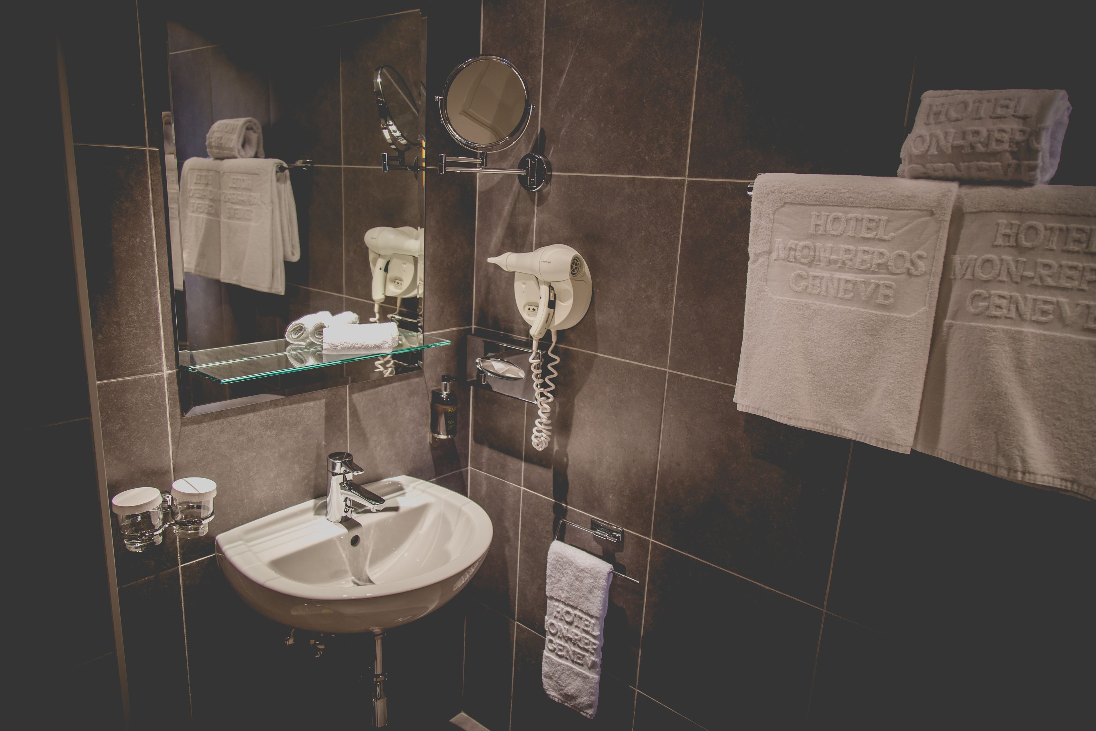 Une salle de bain d’hôtel équipée d’un lavabo, d’un miroir, d’un sèche-cheveux et de serviettes rangées sur un porte-serviettes.