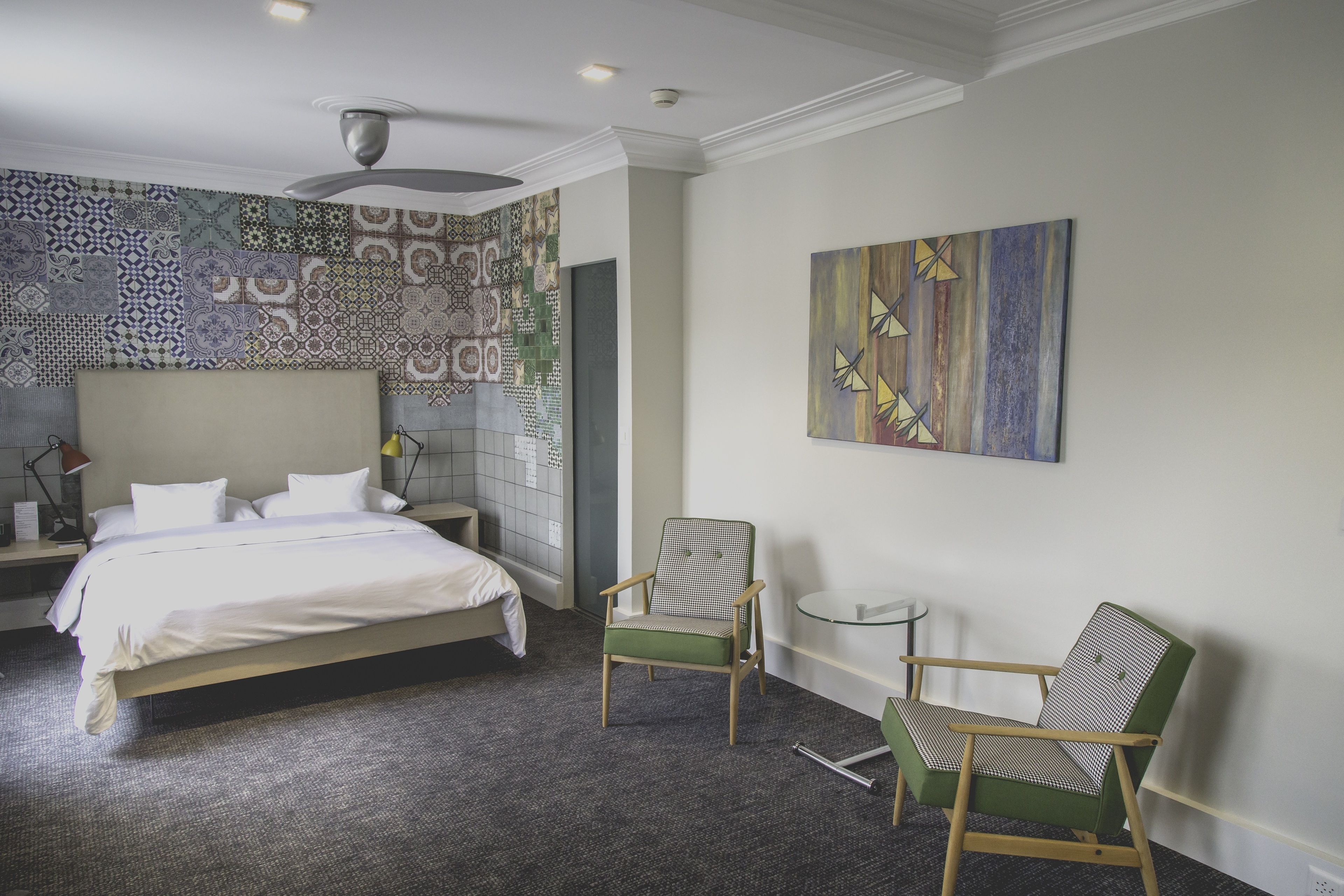 A hotel room with a bed, two chairs, a small table, a ceiling fan, and a patterned wall.