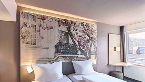 A hotel room with a large bed, a nightstand, a small table, and a window with a view of a building. - B&B Hotel Bonn-City (Bonn)
