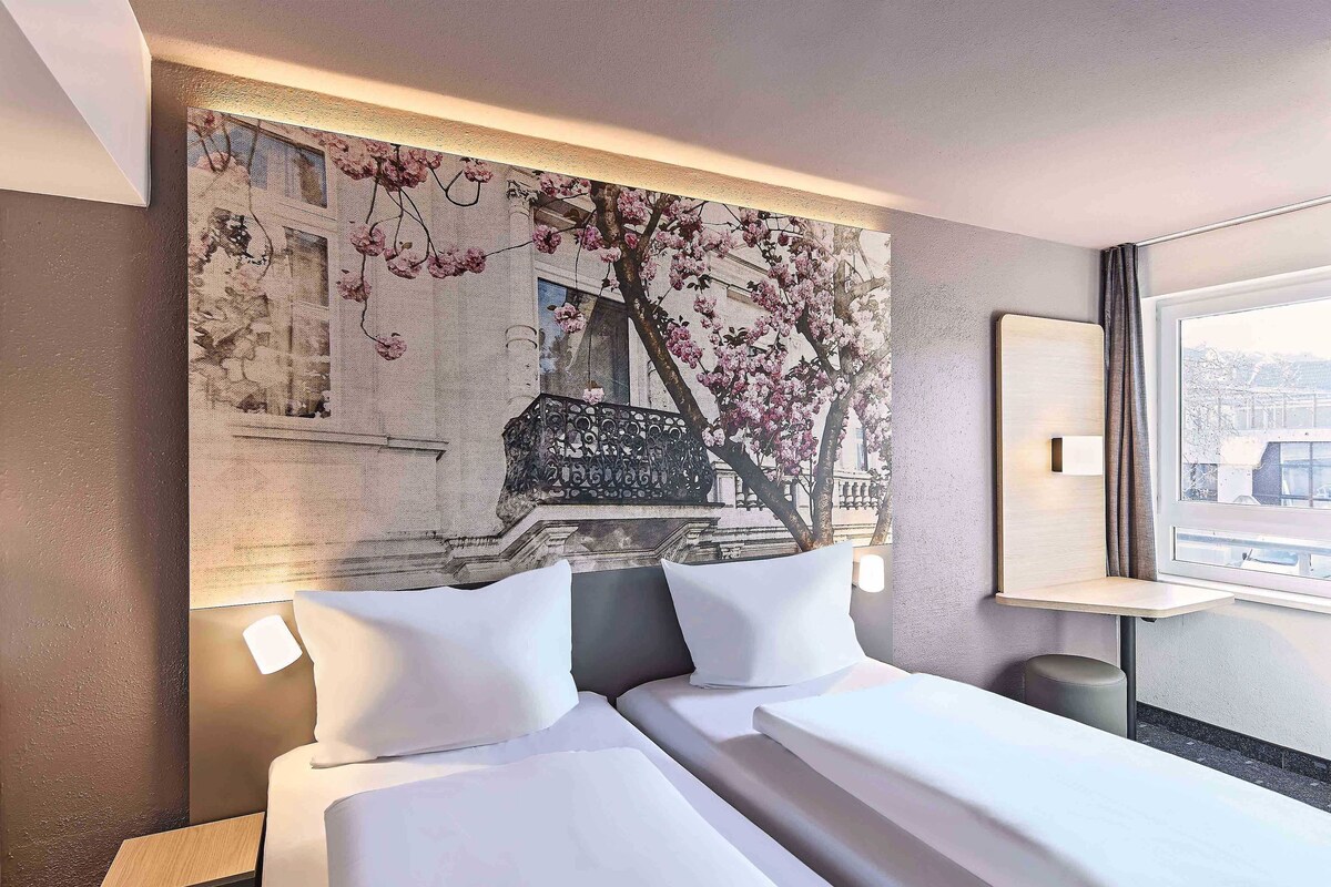 A hotel room with a large bed, a nightstand, a small table, and a window with a view of a building.