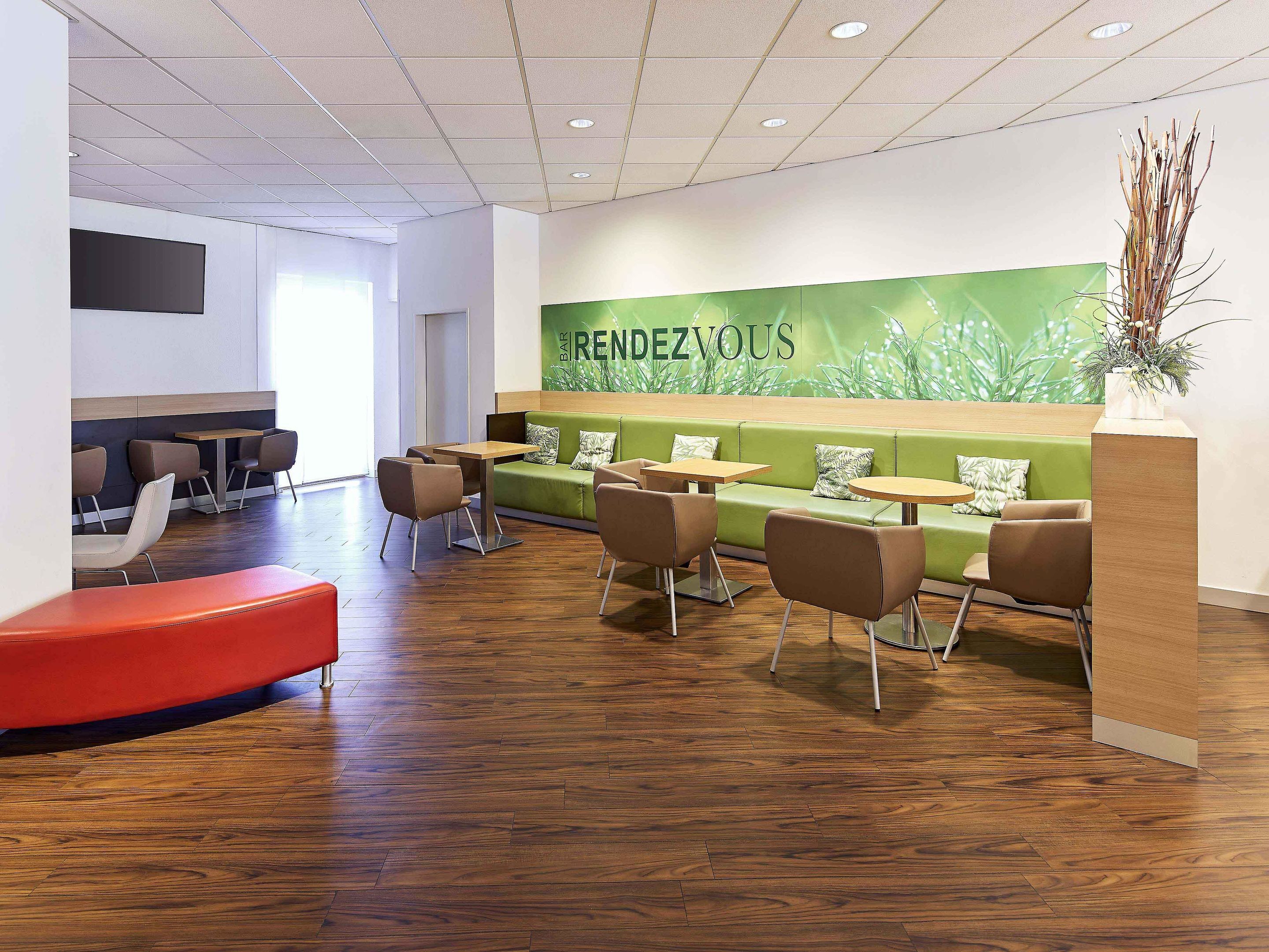 Ein modernes Interieur mit Sitzbereichen, einer Rezeption und einer grünen Wand mit der Aufschrift 'RENDEZVOUS'.