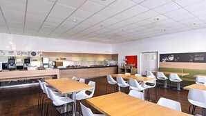 A modern cafe with wooden tables and white chairs, a coffee bar, and a red accent wall. - B&B Hotel Bonn-City (Bonn)