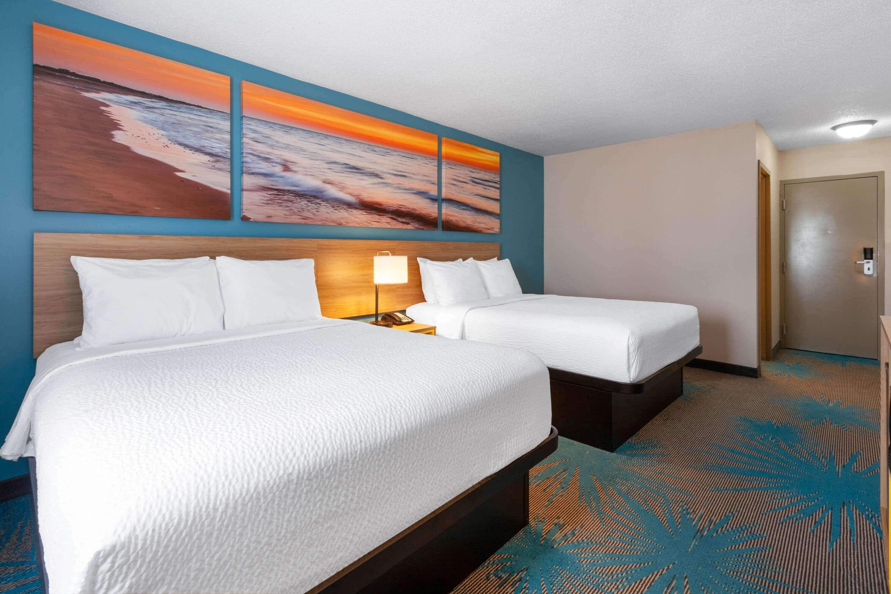A hotel room with two beds, a nightstand, a lamp, and a large wall painting of a sunset over the ocean.