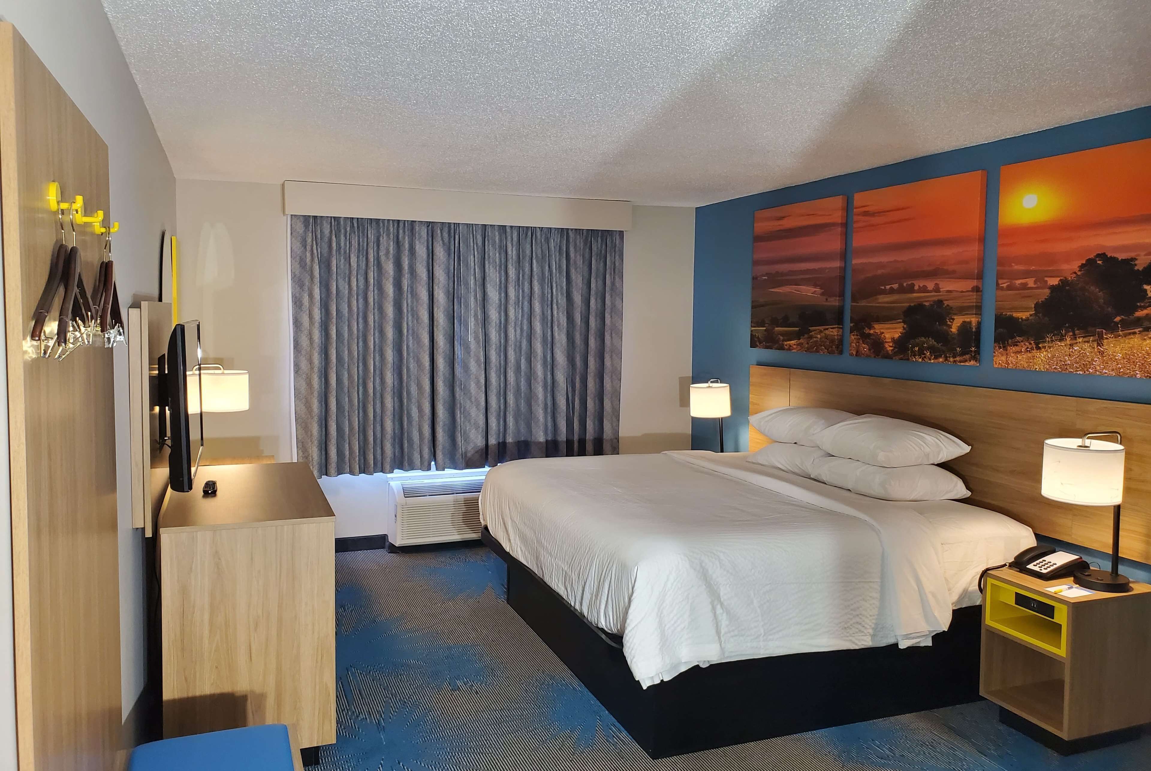 Quarto de hotel com cama, mesa de cabeceira, abajur e uma pintura do pôr do sol.