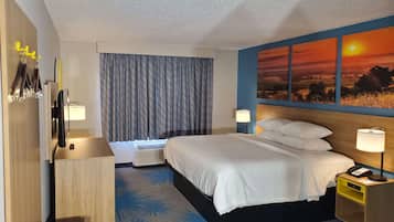 Quarto de hotel com cama, mesa de cabeceira, abajur e uma pintura do pôr do sol.