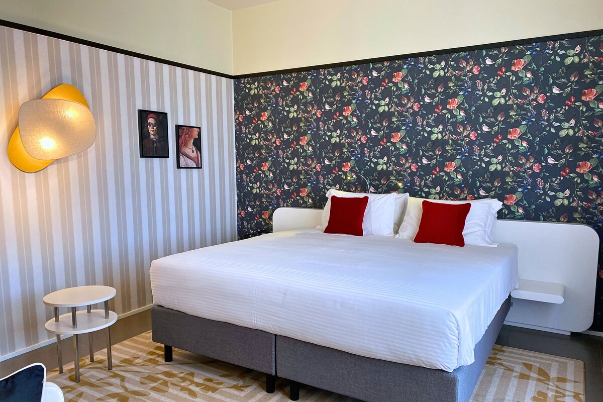 A modern bedroom with a large bed, a small bedside table, a wall-mounted lamp, and a floral wallpaper.