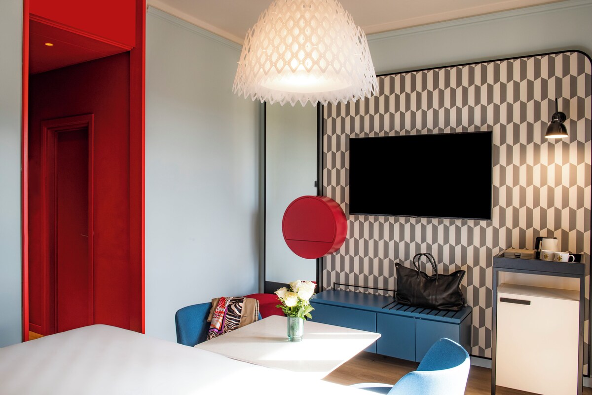 A modern hotel room with a dining area, a flat-screen TV mounted on the wall, a red circular accent piece, and a blue sofa.