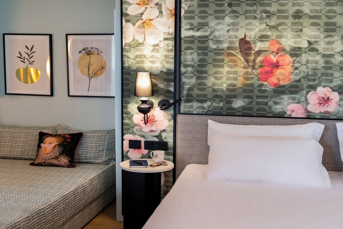 A hotel room with a bed, a sofa, a nightstand with a lamp, and two framed artworks on the wall.