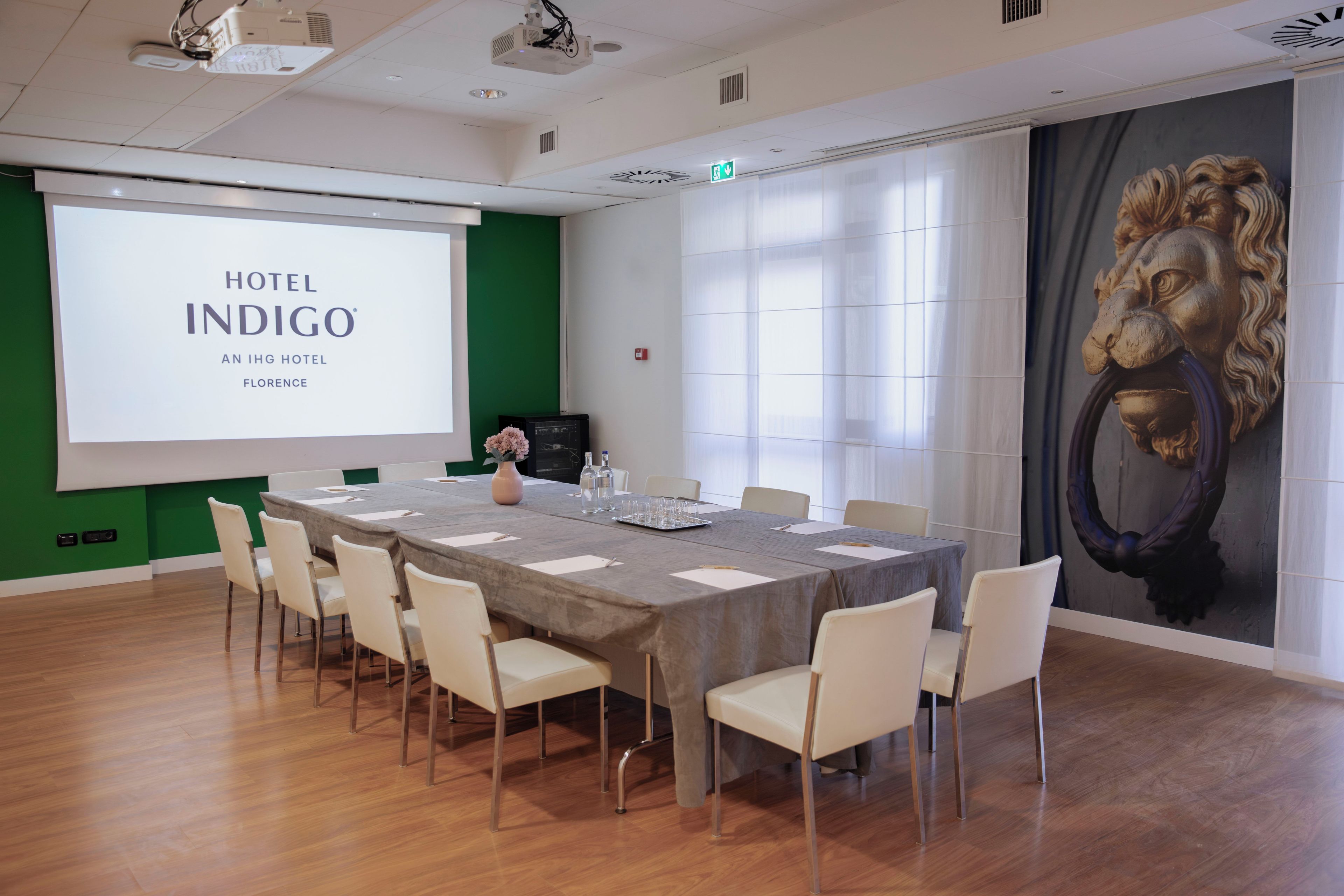 A conference room with a large screen displaying 'HOTEL INDIGO AN IHG HOTEL FLORENCE,' a long table with chairs, and a lion mural on the wall.