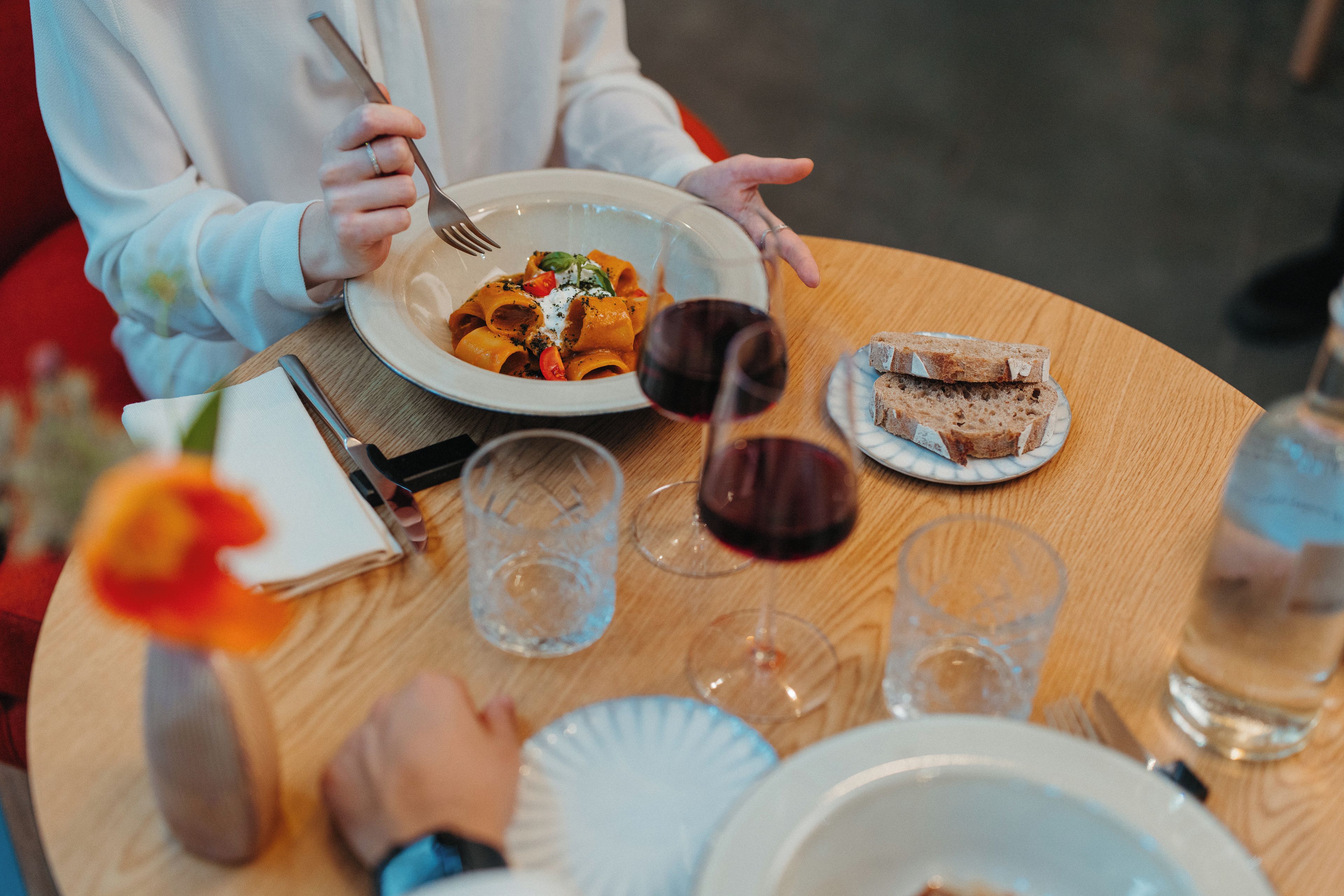 A dining table with a plate of pasta, a glass of red wine, and a slice of bread.