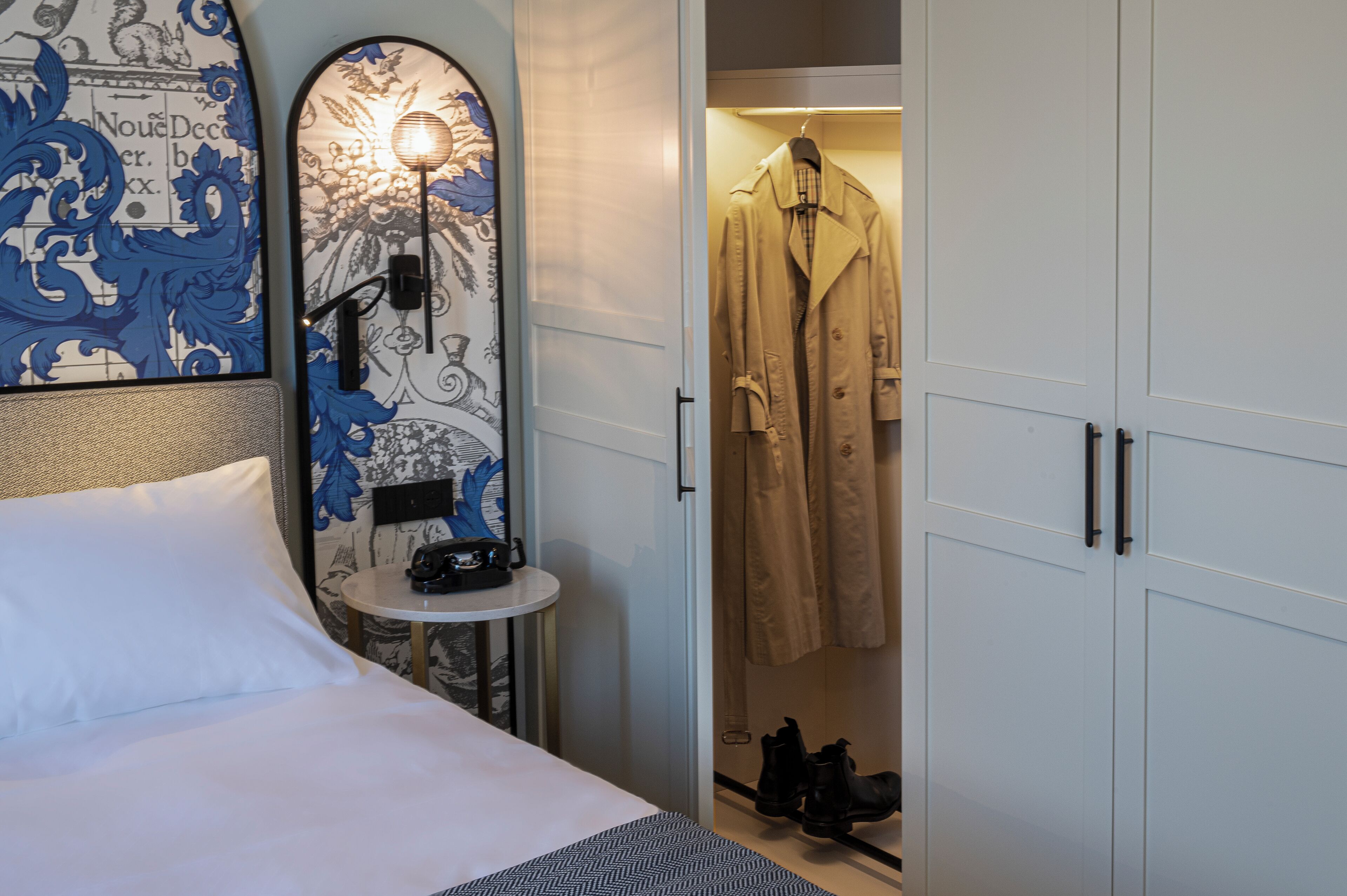 A hotel room with a bed, a small table with a lamp, a coat hanging, and a wardrobe.