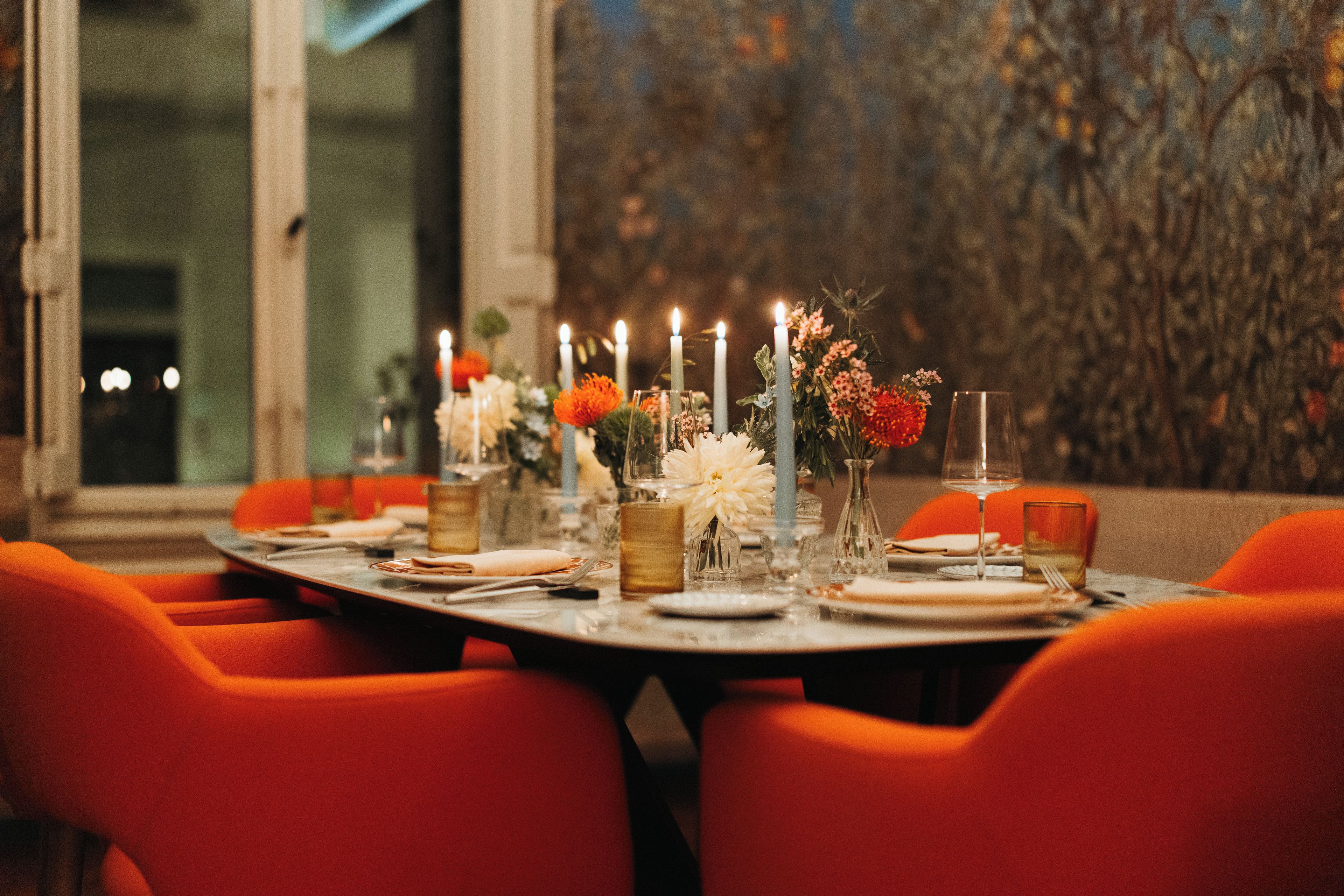 A dining table set with candles, flowers, and glasses, surrounded by orange chairs.