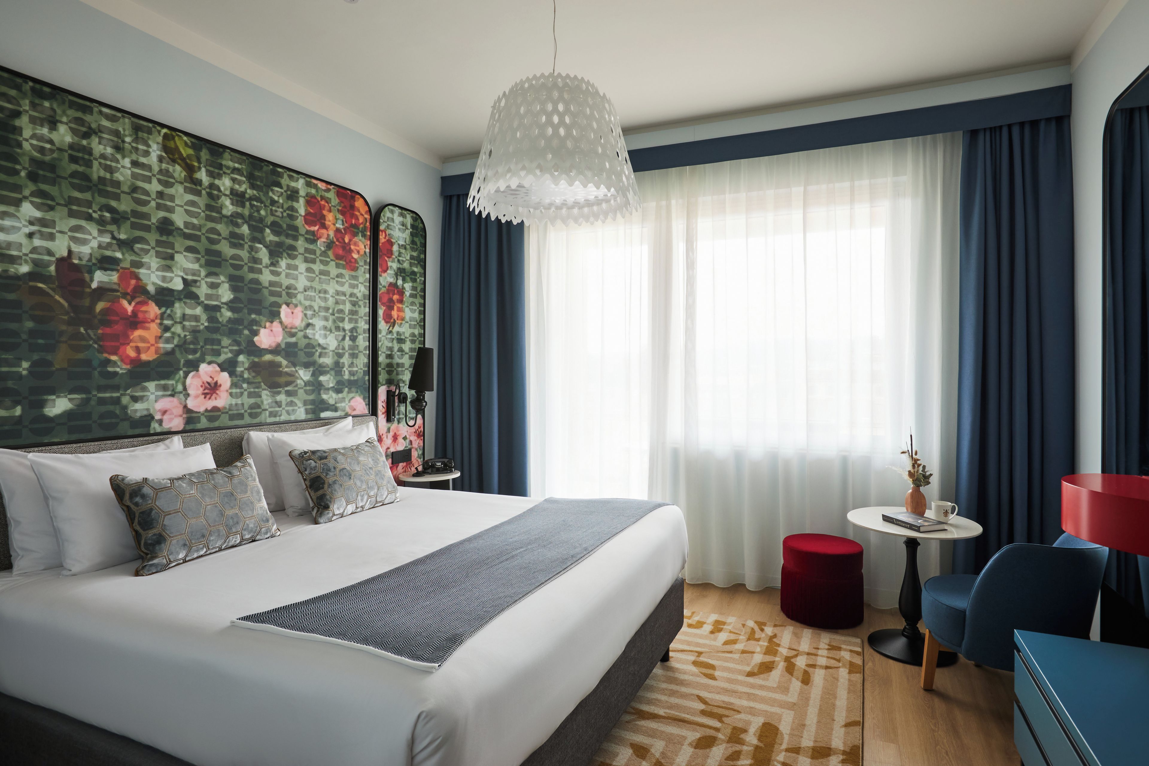 A hotel room with a large bed, a small round table with a lamp, a red ottoman, and a blue chair.