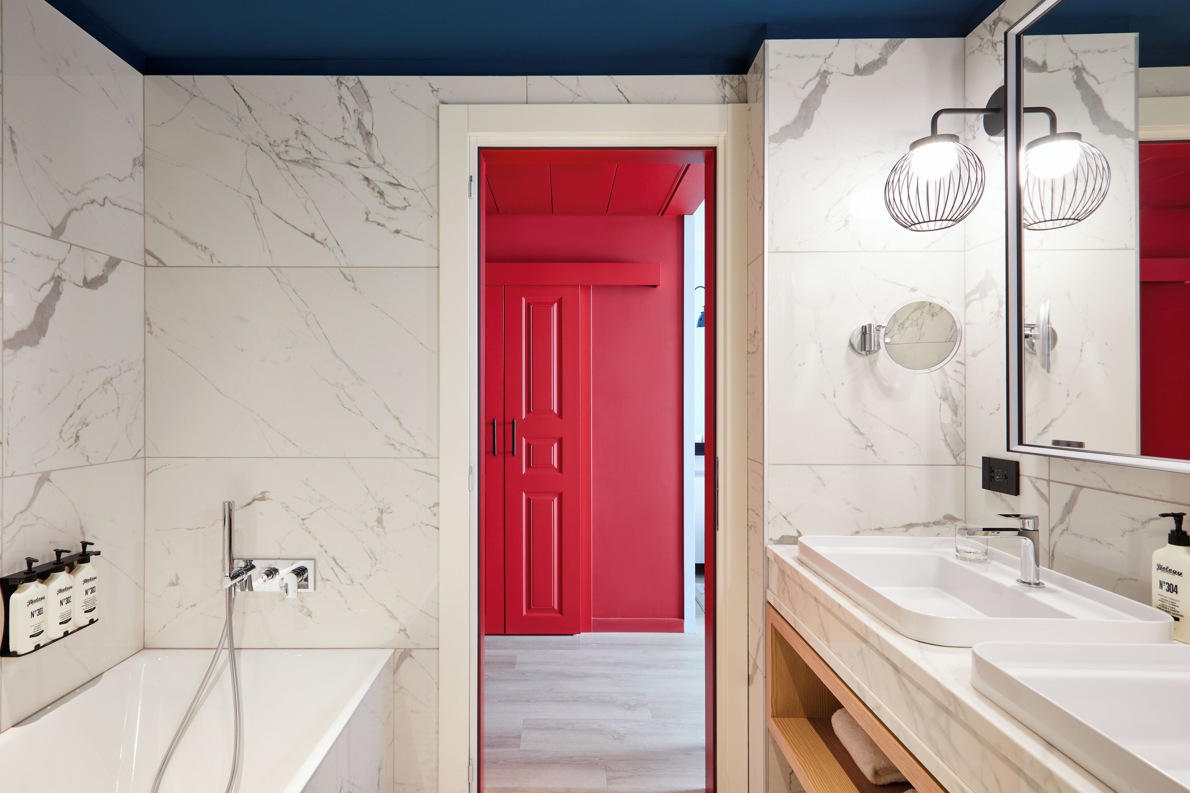 A modern bathroom with a marble wall, a red door, a bathtub, and a vanity with a mirror.