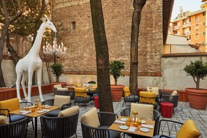 Restaurante con mesas al aire libre, una gran estatua de jirafa blanca y un edificio de ladrillo al fondo.