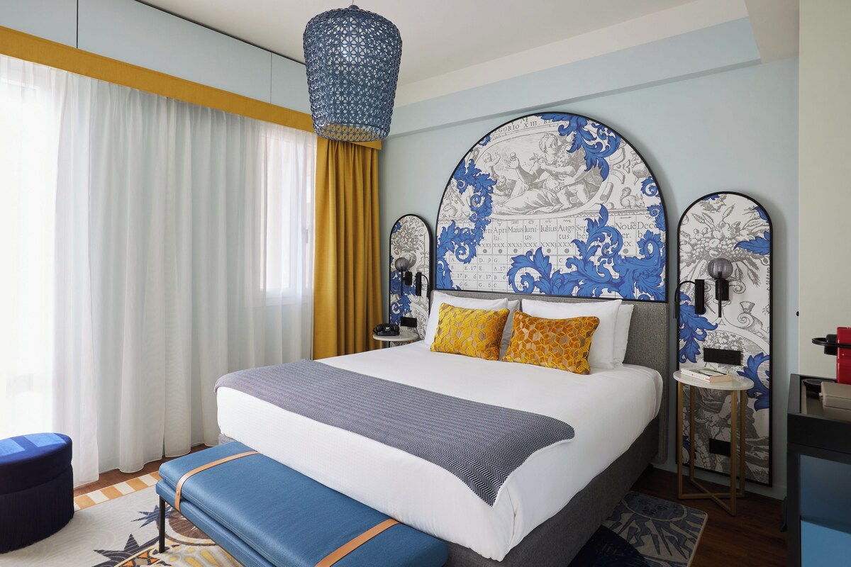 A modern hotel room with a large bed, a bedside table, a nightstand, and a decorative mirror.