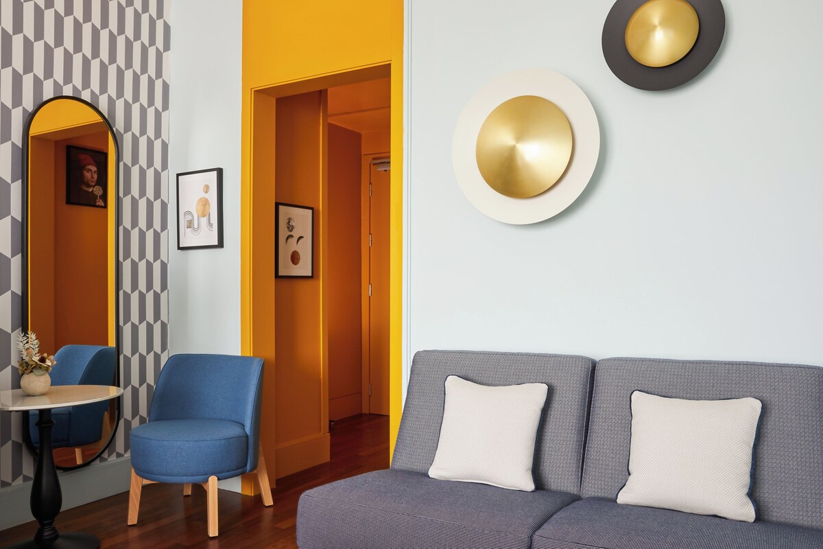 A modern living room with a grey sofa, a blue armchair, and a small table with a vase. There are framed pictures on the wall and a mirror with a geometric pattern.